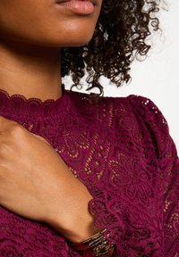 Blusa di pizzo color bordeaux con scollatura a scallop e motivi floreali, abbinata a un bracciale rigido dorato. Dettaglio ravvicinato di spalla e colletto.
