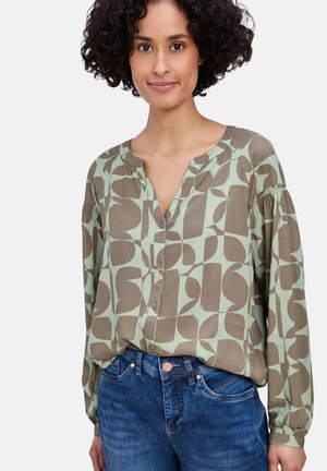 Femme aux cheveux bouclés portant un chemisier boutonné à motifs verts et marron ainsi qu'un jean bleu, debout devant un fond uni.