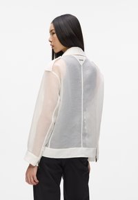 Veste en mesh blanc au design structuré, avec un motif à rayures horizontales, un col et des zips latéraux. Vue de dos montrant un matériau transparent.