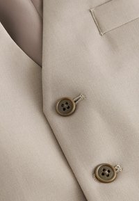 Blazer beige à texture lisse, avec deux boutons marron à quatre trous chacun et un détail de boutonnière cousu.