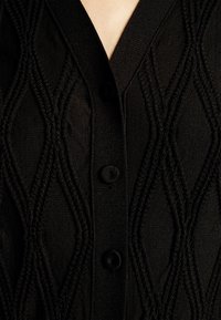 Černý pletený cardigan s diamantovým vzorem, V-krkem a čtyřmi knoflíky vpředu. Texturovaná tkanina dodává designu hloubku.