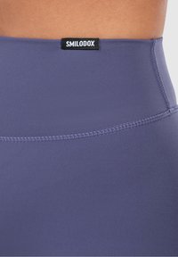Detailní záběr na fialové sportovní legíny s vysokým pasem, viditelným švem a značkovým štítkem "SMILODOX" na pase.