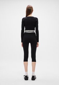 Μαύρη μπλούζα με μακρύ μανίκι και V-κοπή στην πλάτη, συνδυασμένη με μαύρα κολάν 3/4. Διαθέτει ελαστική ζώνη με λογότυπο και μαύρα παπούτσια με άσπρες κάλτσες.