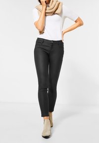 Black skinny jeans with a smooth texture, featuring a mid-rise waist, front pockets, and a close fit, paired with a light top and beige ankle boots.