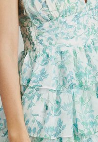 Vestido floral de tela ligera con patrones de hojas verdes y azules, que presenta un cuerpo fruncido y volantes en capas en la falda.
