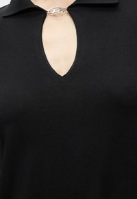 Top en maille noire avec un profond décolleté en V, fermé au col par un fermoir ovale argenté.