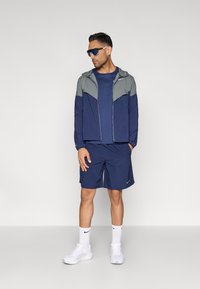 Αθλητική εμφάνιση σε ναυτικό μπλε με ζακέτα με φερμουάρ και γκρι λεπτομέρειες, ταιριαστό t-shirt, σορτς και λευκά αθλητικά παπούτσια με λογότυπο.