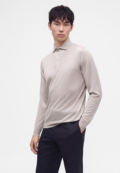Jeune homme aux cheveux courts et noirs portant un polo beige clair à manches longues et un pantalon foncé, debout avec une main dans la poche.
