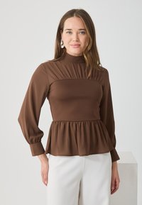 Kobieta z jasnobrązowymi włosami, nosząca srebrne kolczyki, brązową bluzkę z długim rękawem w stylu peplum oraz białe spodnie, stojąca na tle jednolitego tła.