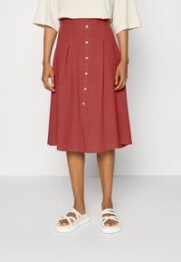 Jupe rouge rouille mi-longue à boutons avec des plis, portée avec un top beige et des sandales plates blanches à lanières sur un fond clair.