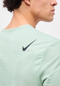 Νέο άτομο που φοράει ανοιχτό πράσινο διατρητό αθλητικό μπλουζάκι με μαύρο λογότυπο Nike στο πάνω μέρος της πλάτης, στραμμένο πλάγια.