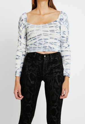 Kobieta w białym i niebieskim crop topie z długim rękawem z efektem tie-dye oraz czarnych, wysokich spodni jeansowych z wzorem węża, stojąca na białym tle.