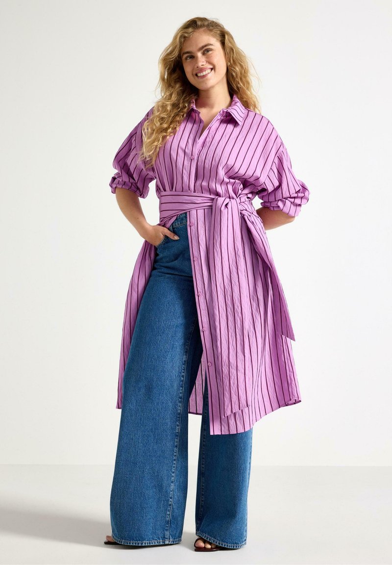 Lilla stribet skjortekjole med korte puffede ærmer og bindebånd i taljen. Kombineret med blå denimbukser med vide ben. Glat tekstur, lukning med knapper.