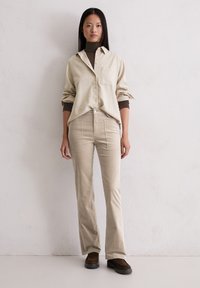 Beige overhemd met knopen en een zak, gecombineerd met lichtbeige uitlopende broek. De outfit heeft een zachte textuur en donkere bruine schoenen.