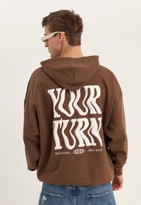 Vīrietis brūnā kapučjakā ar izplūdušiem baltiem burtiem "YOUR TURN" uz muguras, kopā ar ziliem džinsiem un bēšām saulesbrillēm, stāvot ar muguru pret skatītāju.