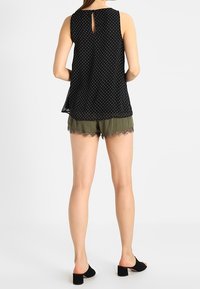 Top senza maniche nero con pois bianchi, dettaglio con apertura a chiave sul retro, abbinato a pantaloni corti verde oliva con bordo in pizzo, indossato con scarpe nere con tacco blocco.
