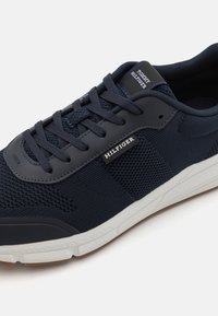 Marineblauwe Tommy Hilfiger sneaker met mesh- en lederen details, witte zool, zwarte veters en "Hilfiger" branding op de zijkant en de tong.