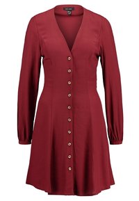 Punainen pitkähihaista mekkoa, jossa on V-pääntie, nappikiinnitys edessä ja laskettu helma. Valmistettu sileästä kankaasta, jossa on hienovarainen kiilto.