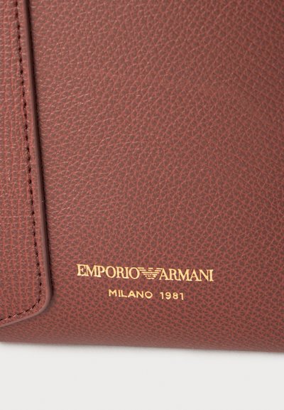 Καφέ δέρμα με υφή και χρυσό λογότυπο Emporio Armani Milano 1981 τυπωμένο κοντά στην ραμμένη άκρη μιας τσάντας ή πορτοφολιού.