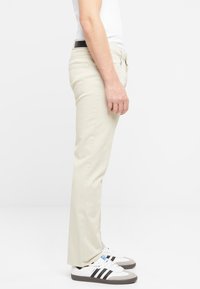 Pantalones rectos beiges de algodón, con bolsillos laterales y traseros, llevados con zapatillas blancas y grises con rayas negras.