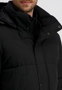 Schwarze Daunenjacke mit strukturiertem Außenmaterial, hohem Kragen und Druckknopf-/Knopfverschlüssen. Das Innere hat ein dezentes gemustertes Futter.