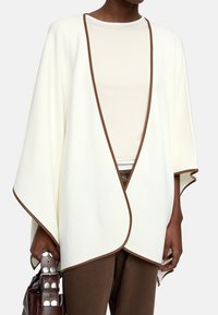 Otwarte na przód, kremowe kardigan z brązowym wykończeniem, wykonane z miękkiego materiału. Posiada szerokie rękawy i luźny krój, w połączeniu z brązową torebką.