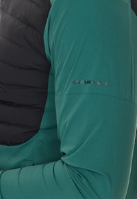 Grön sportjacka med svarta vadderade axlar. Strukturerad tyg på ärmarna, med texten "ENDURANCE" präglad på vänster arm.