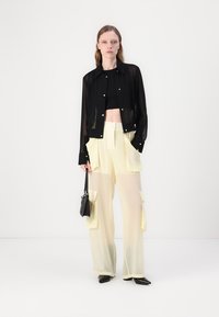 Čierna priesvitná bunda s gombíkmi cez čierny crop top, skombinovaná so svetlo žltými cargo nohavicami s veľkými vreckami a čiernymi topánkami.