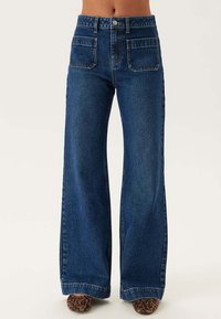 Mörkblå denimjeans med vida ben som har hög midja, fyra fickor och en klassisk knapp- och dragkedjestängning. Strukturerad tyg med en slät yta.