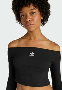 Czarny prążkowany crop top z odsłoniętymi ramionami, z długimi rękawami i białym logo Adidas na piersi, podkreślający dopasowany fason i gładką teksturę.