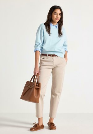 Giovane donna in piedi con borsa marrone, indossa un maglione azzurro chiaro sopra una camicia a righe, pantaloni beige e scarpe di pelle marrone su sfondo uniforme.