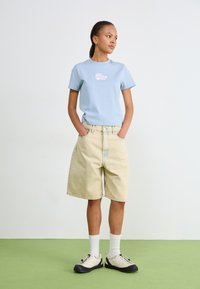 Ljusblå bomullströja med en rosa snäcka grafik, i kombination med beige denimshorts. Modellen har vita strumpor och beiga skor med svarta sulor.