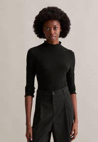 Schwarzes, geripptes Langarmoberteil mit gerafftem Ausschnitt, kombiniert mit dunkel gestreiften Hosen, die texturierte Stoffe und eine figurbetonte Passform zeigen.