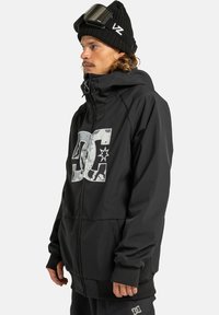 Uomo che indossa una giacca con cappuccio nera con il logo DC bianco, un berretto nero con occhiali da sci e pantaloni neri con il logo DC, rivolto a sinistra.