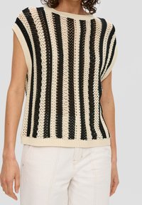 Beżowy, bezrękawnik dzianinowy z pionowymi czarnymi paskami, noszony z kremowymi spodniami, widoczny od ramion do bioder.