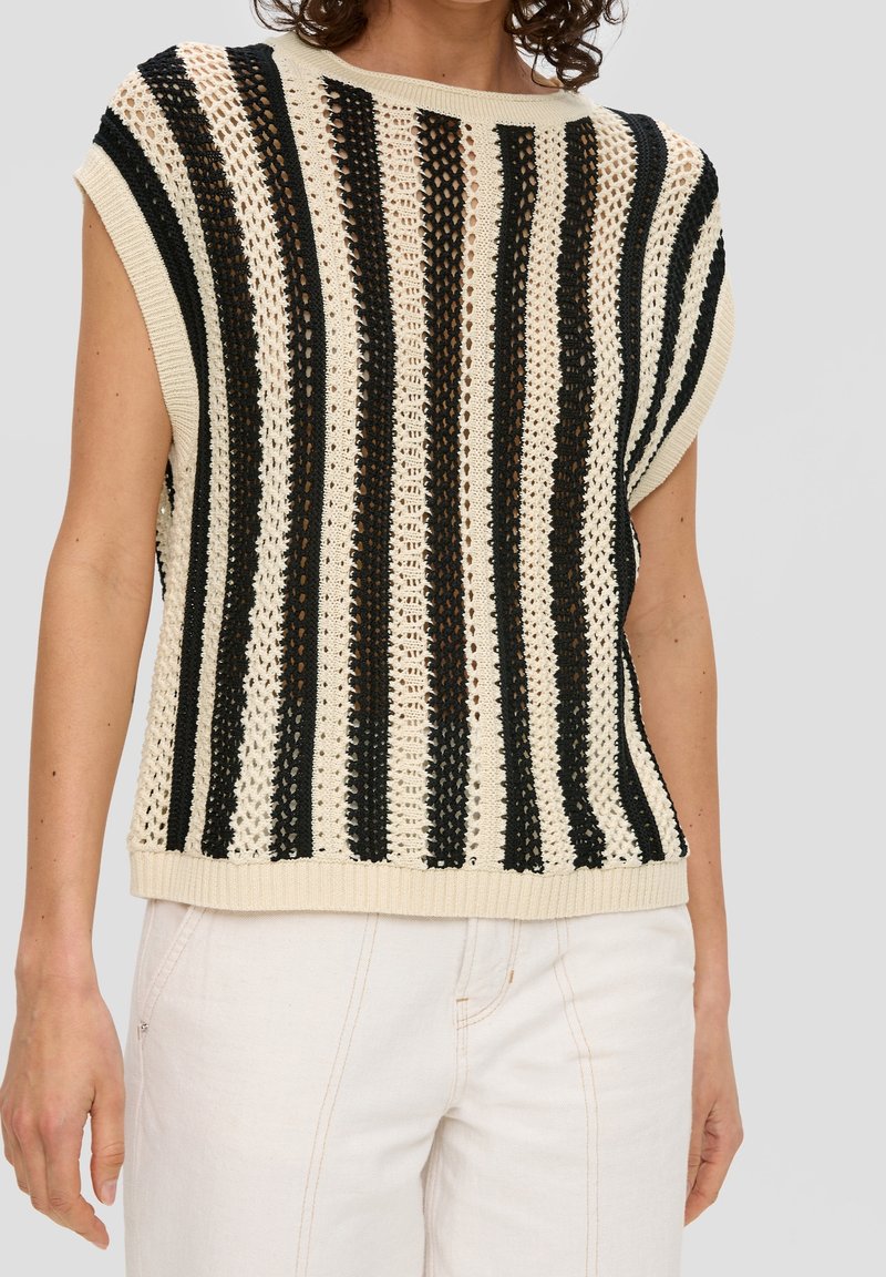 Beżowy, bezrękawnik dzianinowy z pionowymi czarnymi paskami, noszony z kremowymi spodniami, widoczny od ramion do bioder.