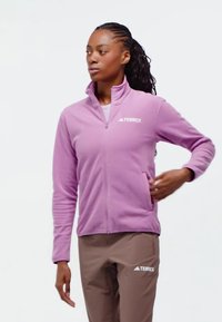 Helllila Fleecejacke mit hohem Kragen, Frontreißverschluss und zwei seitlichen Taschen. Auf der linken Brust ist das 'TERREX'-Logo in Weiß angebracht.
