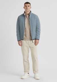 Himmelblau gesteppte Jacke mit Stehkragen, Frontreißverschluss, getragen über einem beigen Rundhalsausschnitt-Shirt und beigen Cordhosen; weiße Sneakers.