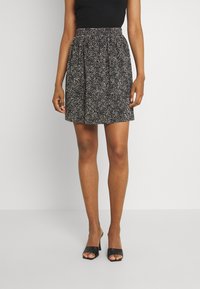 Jupe noire avec de petites taches blanches, taille froncée, design évasé, longueur mi-cuisse, associée à des talons ouverts noirs.