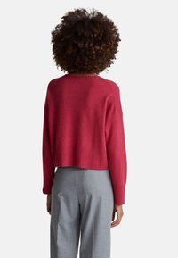 Krótki sweter w głębokim różowym kolorze, dzianinowa faktura, z prążkowanym dołem i mankietami. W zestawie z szarymi, dopasowanymi spodniami, widok od tyłu.