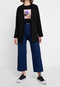 Černý dvouřadý blazer přes černé tričko s grafickým potiskem psa, spárovaný s modrými džíny s vysokým pasem a širokými nohavicemi a černými teniskami.