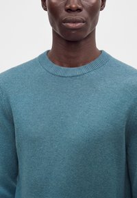 Gros plan d’une personne portant un pull en tricot bleu à col rond sur un fond clair uni, montrant uniquement le cou et les épaules.