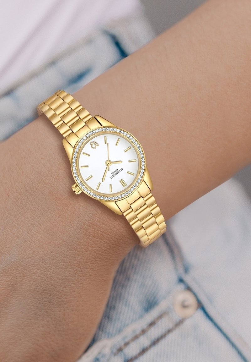 Kuldne randmoots, millel on ümmargune valge kellase, kuldsed ajalised markerid ja sädelev kristallist äär, millel on kuldne lingikett.