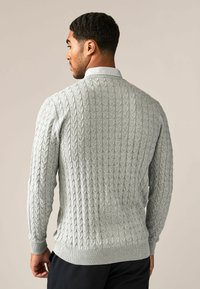 Homme portant un pull en maille légère gris clair sur une chemise à col à carreaux, vu de dos devant un fond uni.