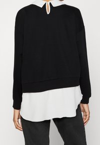 Felpa nera con colletto bianco, caratterizzata da un'apertura a chiave nella parte posteriore e orlo a coste. Indossata sopra una camicia bianca e abbinata a pantaloni scuri.
