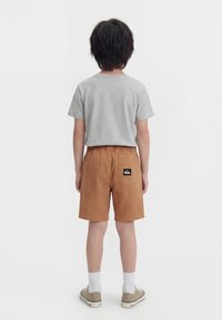 Παιδί με κοντά σκούρα μαλλιά στέκεται με την πλάτη στραμμένη φορώντας ένα ανοιχτό γκρι μπλούζα, καφέ σορτς με λογότυπο στην πίσω τσέπη, λευκές κάλτσες και μπεζ αθλητικά παπούτσια.