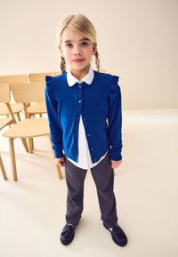 Cardigan blu lavorato a maglia con dettagli arricciati, chiusura con bottoni, indossato sopra una camicia bianca con colletto e pantaloni grigi. Scarpe nere con dettaglio a fiocco.