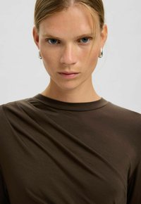 Femme aux yeux bleus, cheveux clairs attachés en arrière, portant des boucles d'oreilles argentées en anneau et un haut marron foncé, regardant directement la caméra.