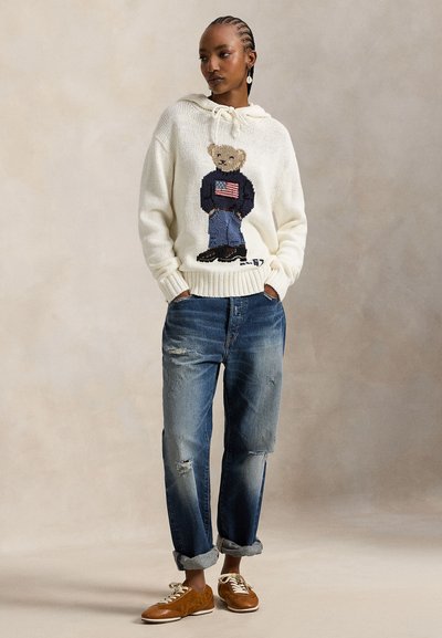 Κρεμ πλεκτό πουλόβερ με σχέδιο αρκούδας και αμερικανική σημαία, συνδυασμένο με σκισμένο μπλε τζιν και καφέ αθλητικά παπούτσια με λευκά κορδόνια.