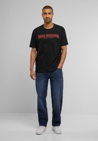 Camiseta de algodón negra con un logo rojo de "True Religion", combinada con jeans de mezclilla azul y zapatillas blancas, de pie contra un fondo neutro.